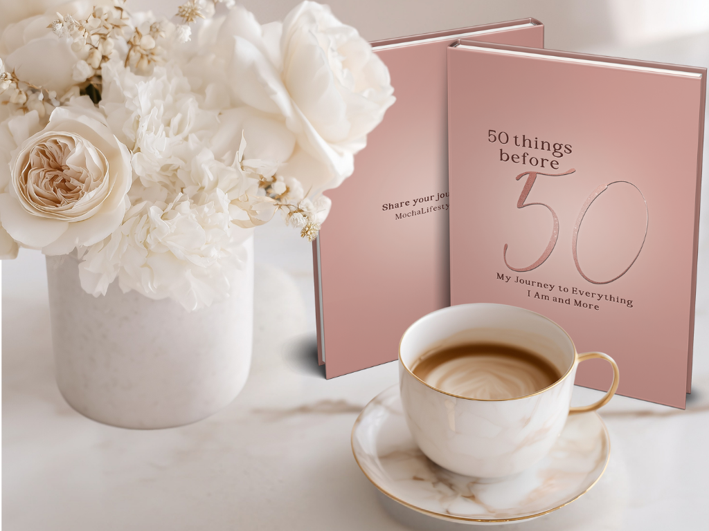 Blush colored journal titled '50 Things Before 50' with a cup of coffee and flowers on a marble surface
