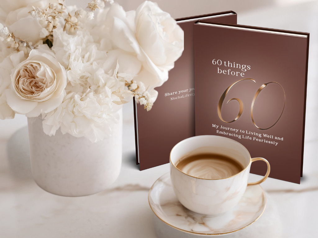 Cup of coffee with a journal titled '60 things before 60' and white flowers on a table.